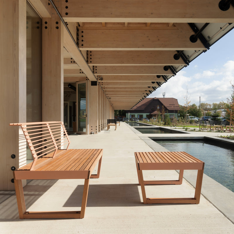 Egoes Barka Public Benches, crafted from Jatoba wood, line a concrete walkway beneath an exposed beam structure. The path, adorned with street furniture, runs alongside a reflective pool with trees, a house, and cloudy sky in the background.