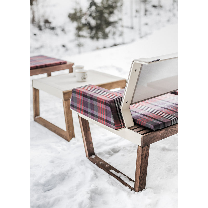 In this cozy outdoor scene, a Barka chair and tables by Egoe rest on a snowy surface. They feature plaid cushions and pillows, accompanied by a cup and saucer. Snow-covered trees create a serene backdrop, highlighting the exquisite design of this outdoor furniture collection.