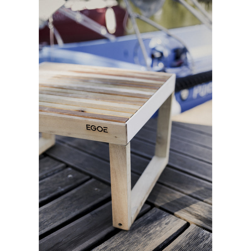 The Barka Stool from Egoe, featuring a striped design, sits elegantly on a wooden deck with part of a blue boat and blurred outdoor scenery enhancing this exquisite outdoor furniture.
