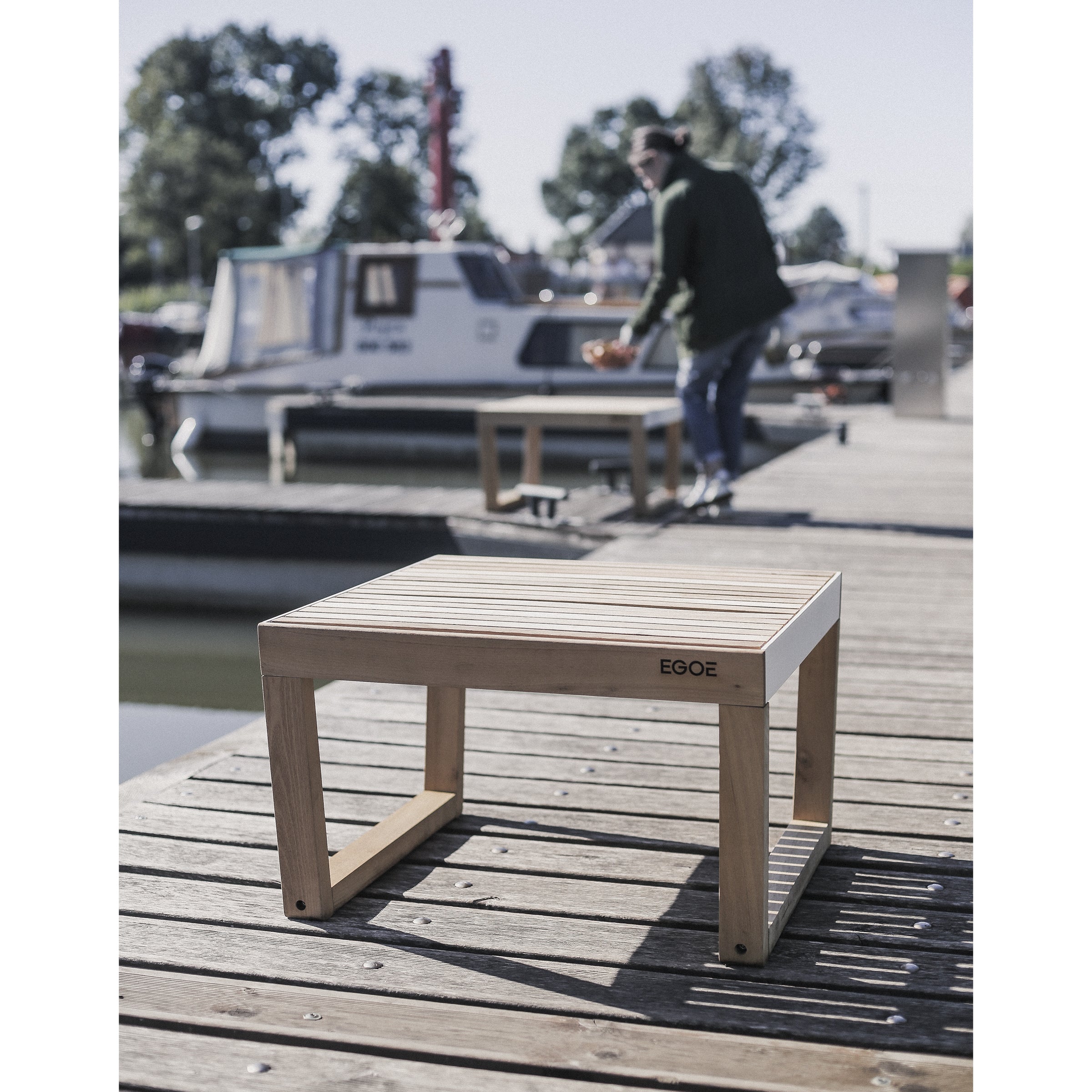 The Barka Stool by Egoe, crafted from exotic wood with EQOE engraved, is showcased on a dock beside water. A person and boat are visible, framed by trees and sky in a serene outdoor setting.