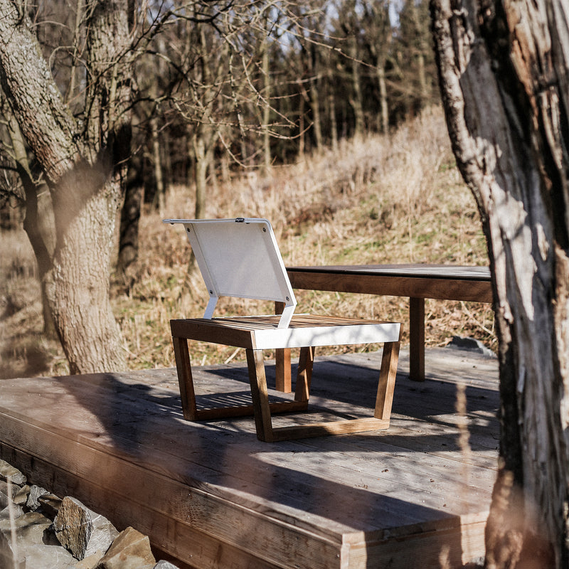 A wooden deck in a forest showcases two Egoe Barka Chairs with minimalist white seats, set against bare trees and dried foliage. Natural light enhances the serene and rustic ambiance, highlighting the beauty of this outdoor furniture.