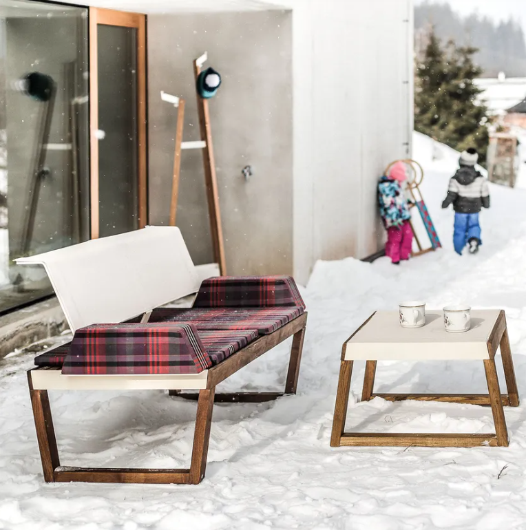 The snowy patio displays Egoes Barka Stool with a plaid cushion, accompanied by a matching table holding two mugs. In the background, a child in winter attire pulls a sled, and reflections and ski poles are visible by the glass door.