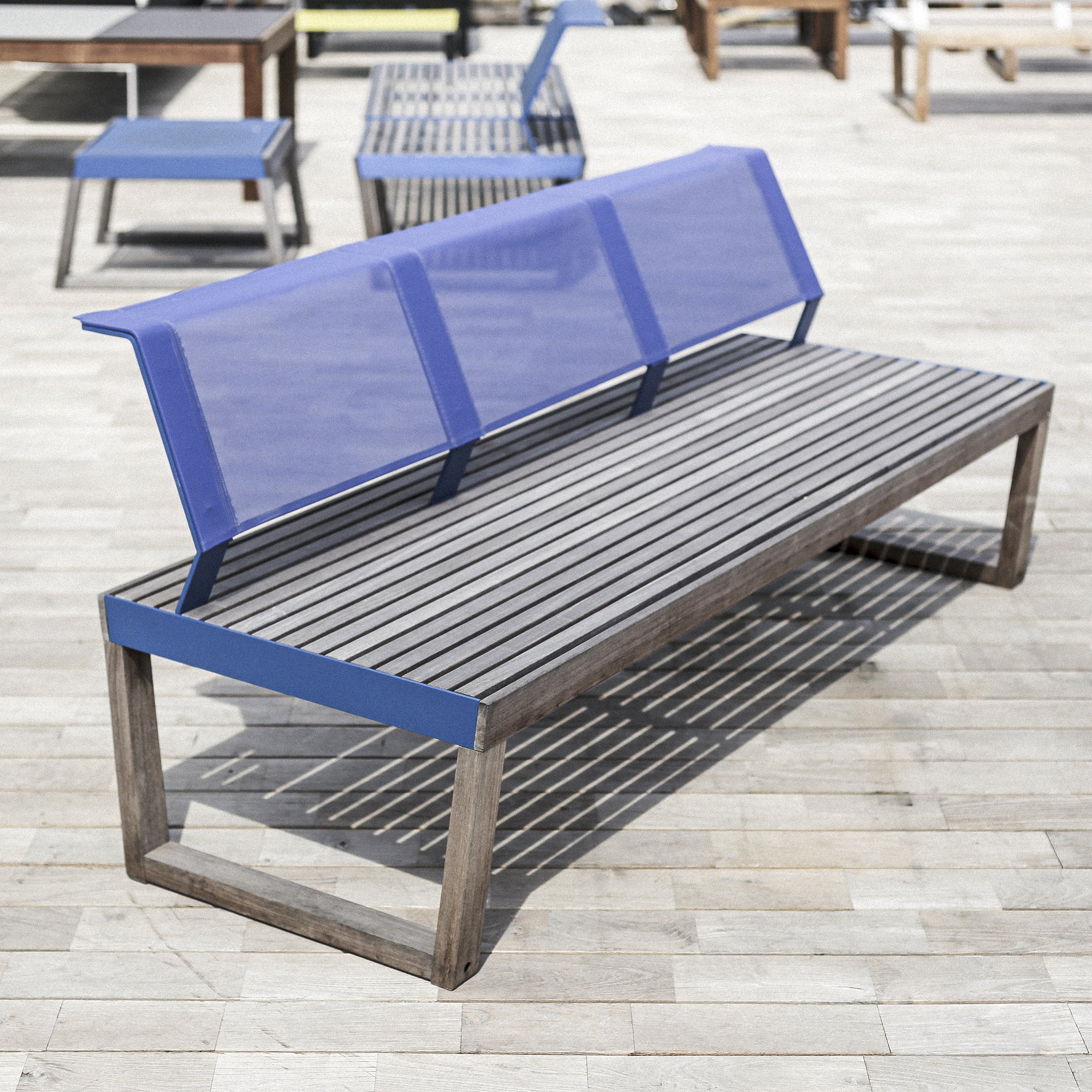 The Barka Bench by Egoe is a modern outdoor piece with a sleek, angular design, featuring a slatted seat made from exotic wood and a blue, semi-transparent backrest. It complements stylish patio furniture when placed on a wooden deck.