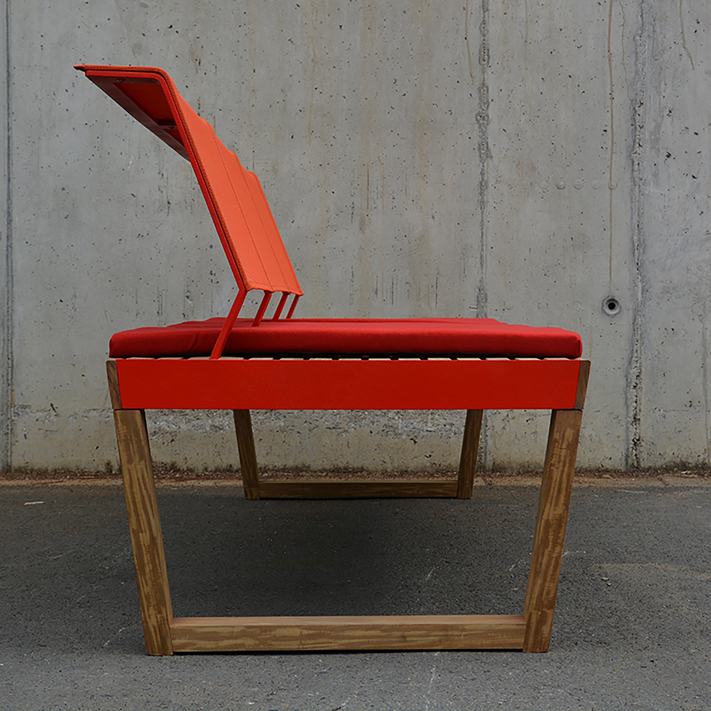 A side view of the Barka Bench by Egoe showcases its exotic wood base and bright red seat with a partially angled backrest. This modern outdoor piece stands out against a textured concrete wall on a gray pavement.