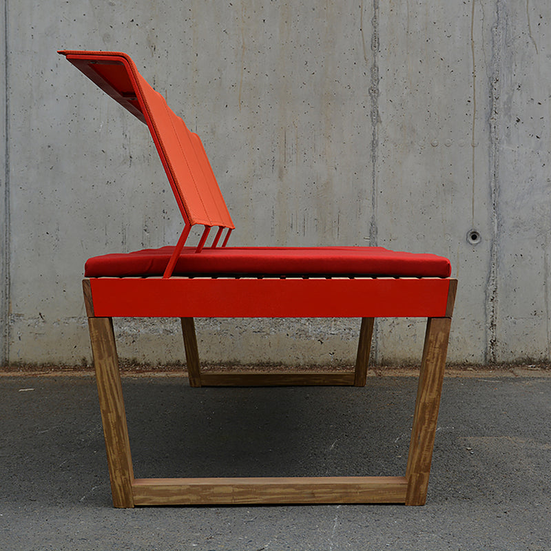 A side view of the Barka Bench by Egoe showcases its exotic wood base and bright red seat with a partially angled backrest. This modern outdoor piece stands out against a textured concrete wall on a gray pavement.