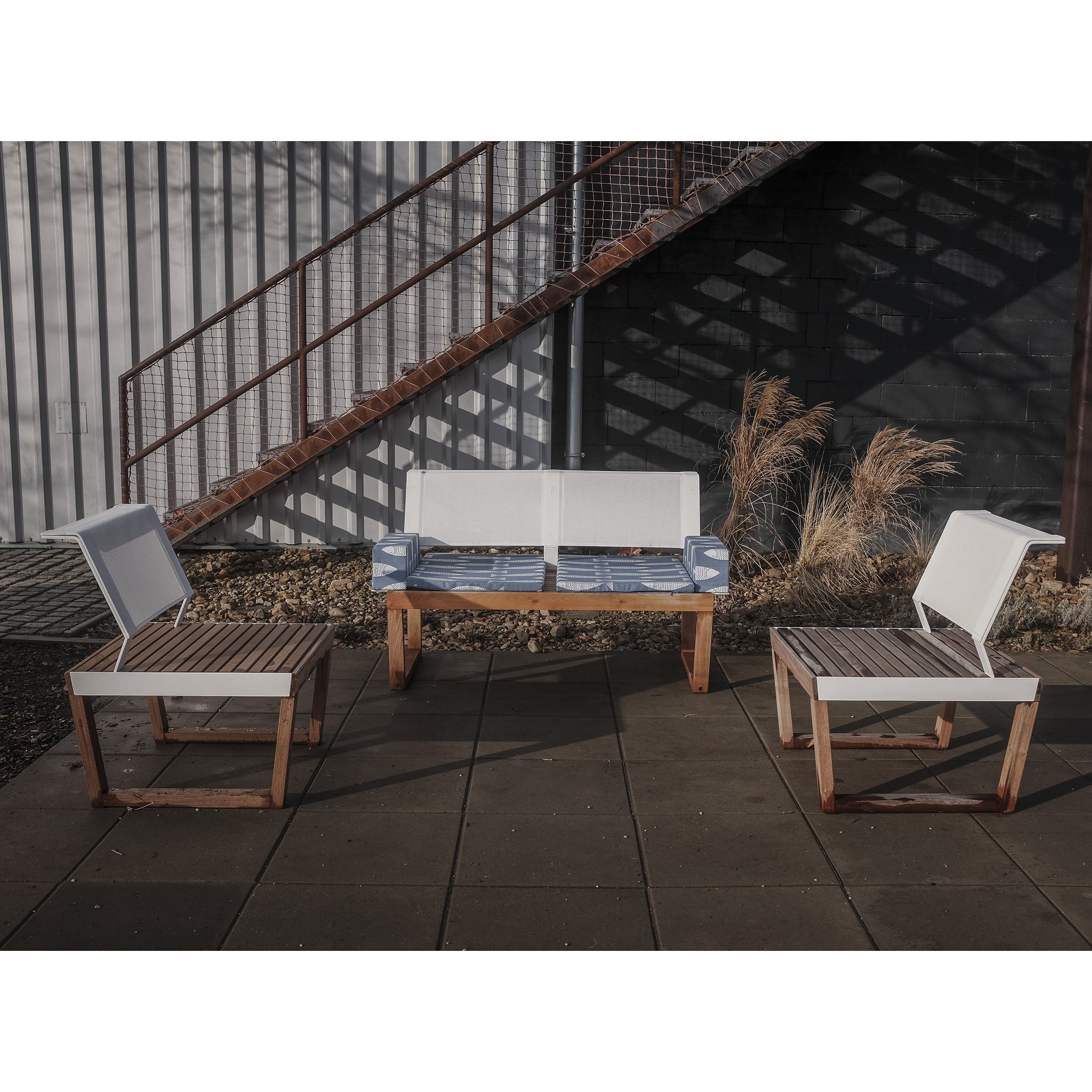 Outdoor seating area features Egoes Barka collection with two chairs and a bench on a tiled patio. The furniture boasts light cushions and exotic wood frames. A metal staircase and ornamental grasses cast shadows on a corrugated wall in the background.