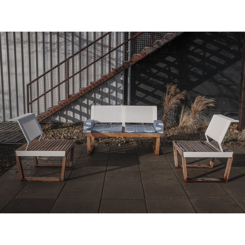 Outdoor seating area features Egoes Barka collection with two chairs and a bench on a tiled patio. The furniture boasts light cushions and exotic wood frames. A metal staircase and ornamental grasses cast shadows on a corrugated wall in the background.