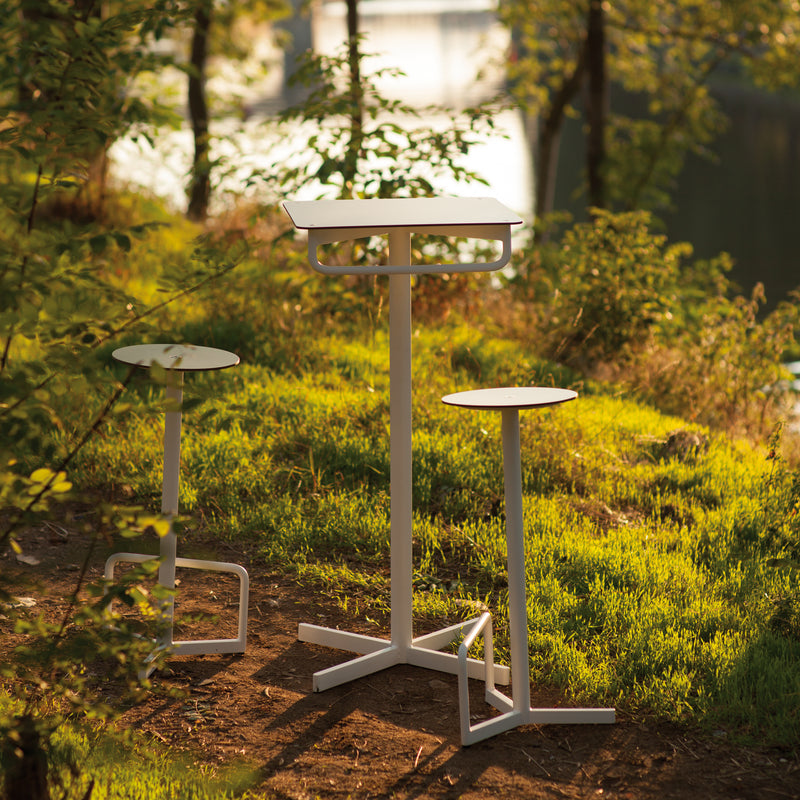 The Egoe BISTROT BAR Bar Table, paired with two modern outdoor stools, creates a chic bar set in a grassy lakeside clearing. The sleek, minimalist white design contrasts with nature, evoking the serene ambiance of elegant restaurant furniture.