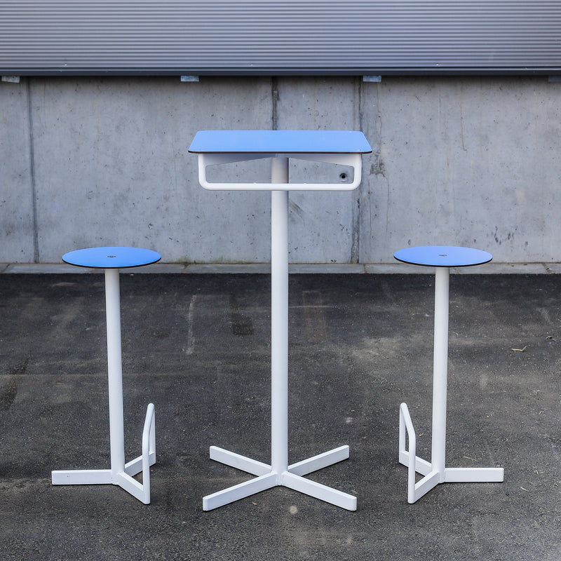 The chic BISTROT BAR Bar Table by Egoe includes two modern metal stools in blue and white, perfectly matching the tall table with a rectangular top. Ideal as restaurant furniture, this sleek set is elegantly displayed on a gray concrete surface against a metal wall backdrop.