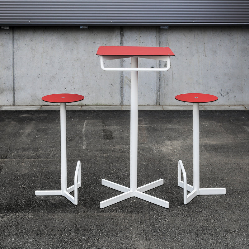 A modern BISTROT BAR set from Egoe featuring two red stools and a matching high table with sleek white metal legs, all stylishly arranged on gray pavement against a concrete wall beneath a metal shutter.