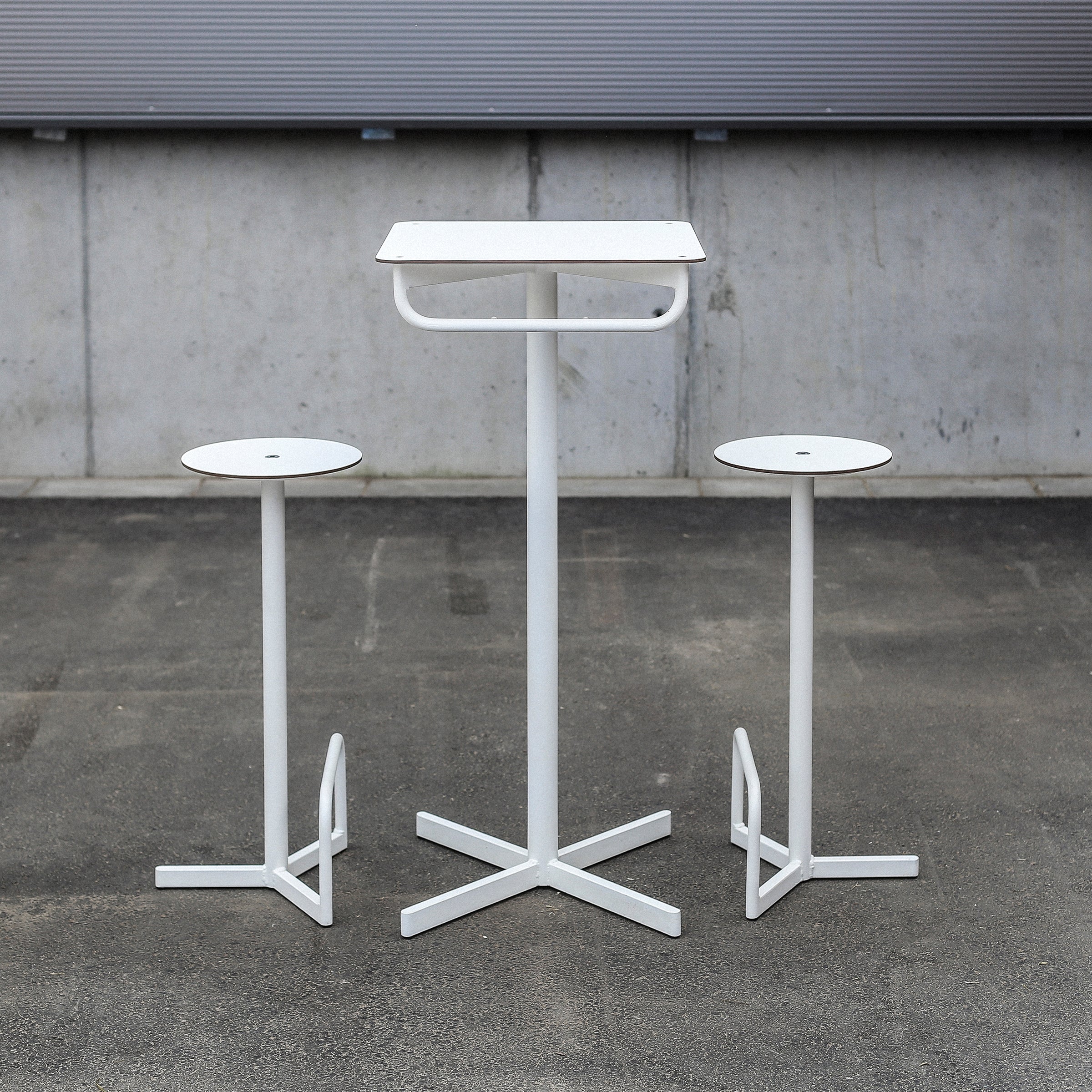 The BISTROT BAR Bar Table by Egoe features a minimalist design with a square white metal table and two matching stools with circular tops, set against a concrete wall and asphalt ground, creating an industrial restaurant furniture vibe.