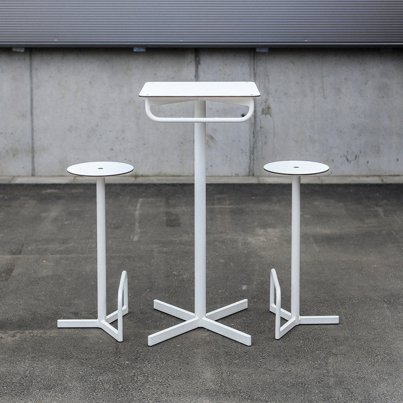 The BISTROT BAR Bar Table by Egoe features a minimalist design with a square white metal table and two matching stools with circular tops, set against a concrete wall and asphalt ground, creating an industrial restaurant furniture vibe.