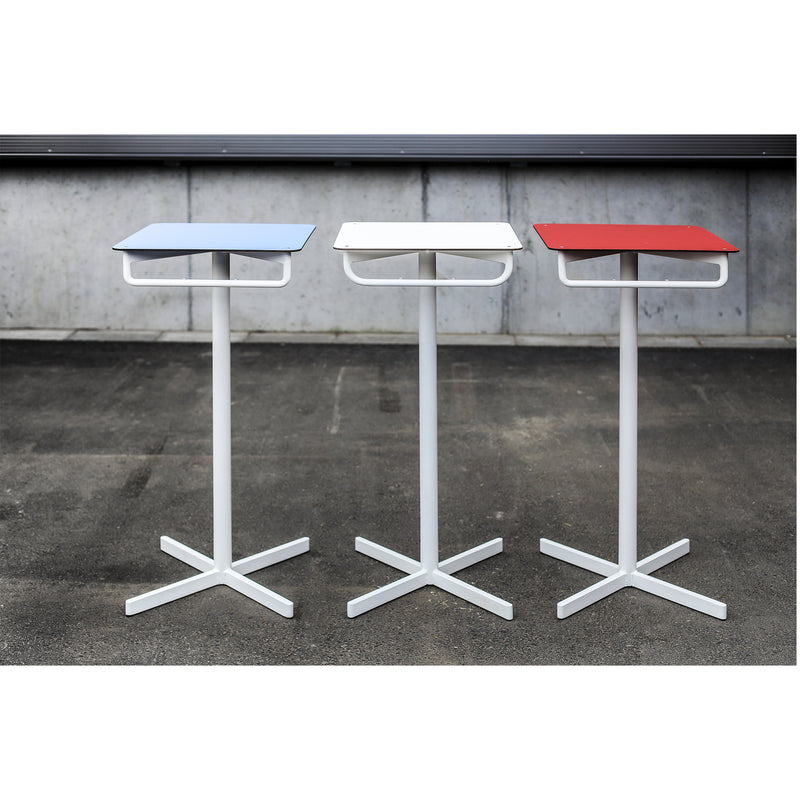 Three BISTROT BAR tables by Egoe stand side by side, creating a chic bar set on a paved surface. Each table features a square top in light blue, white, or red, supported by a white pedestal base with four legs. A concrete wall beautifully complements this stylish restaurant furniture setup.