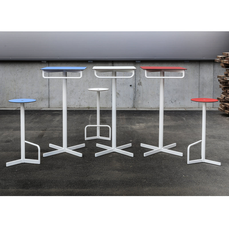 Five Egoe BISTROT BAR tables, featuring tall white metal bases and vibrant red and blue round tops, are arranged in an outdoor industrial setting with a concrete wall and stacked wooden pallets, perfect for a chic bar vibe.