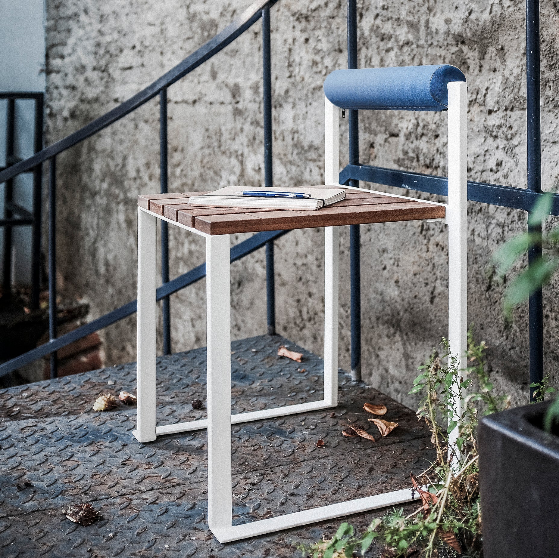The Bistrot Stool with Backrest from Egoe features a seat made of tropical wood and a white zinc-plated steel frame, complemented by a blue cylindrical backrest cushion. Its located outdoors near a stone wall and black metal railing, holding a notebook and pen among scattered leaves.