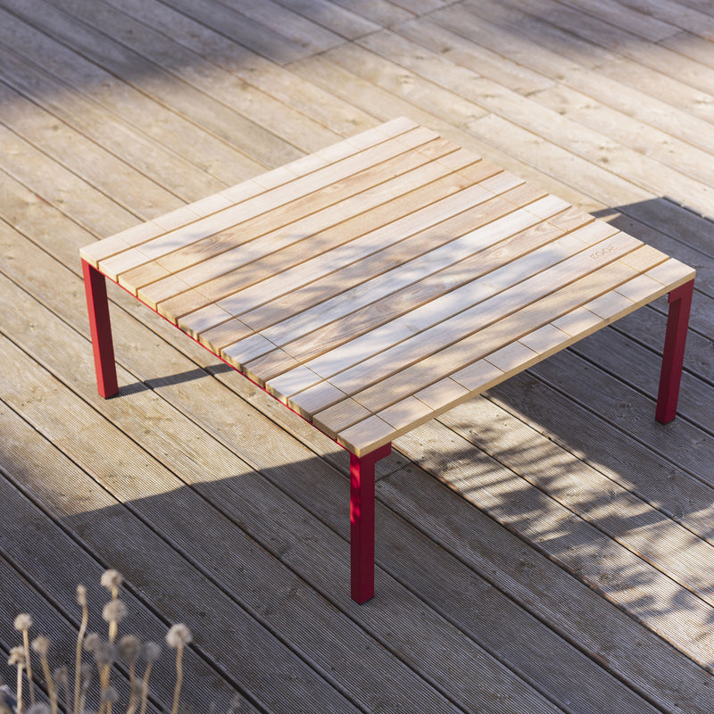 The Bistrot Table by Egoe boasts a slatted tropical wood top and vibrant red zinc-plated steel legs. Perched on a sunlit deck, it beautifully captures artistic branch shadows.