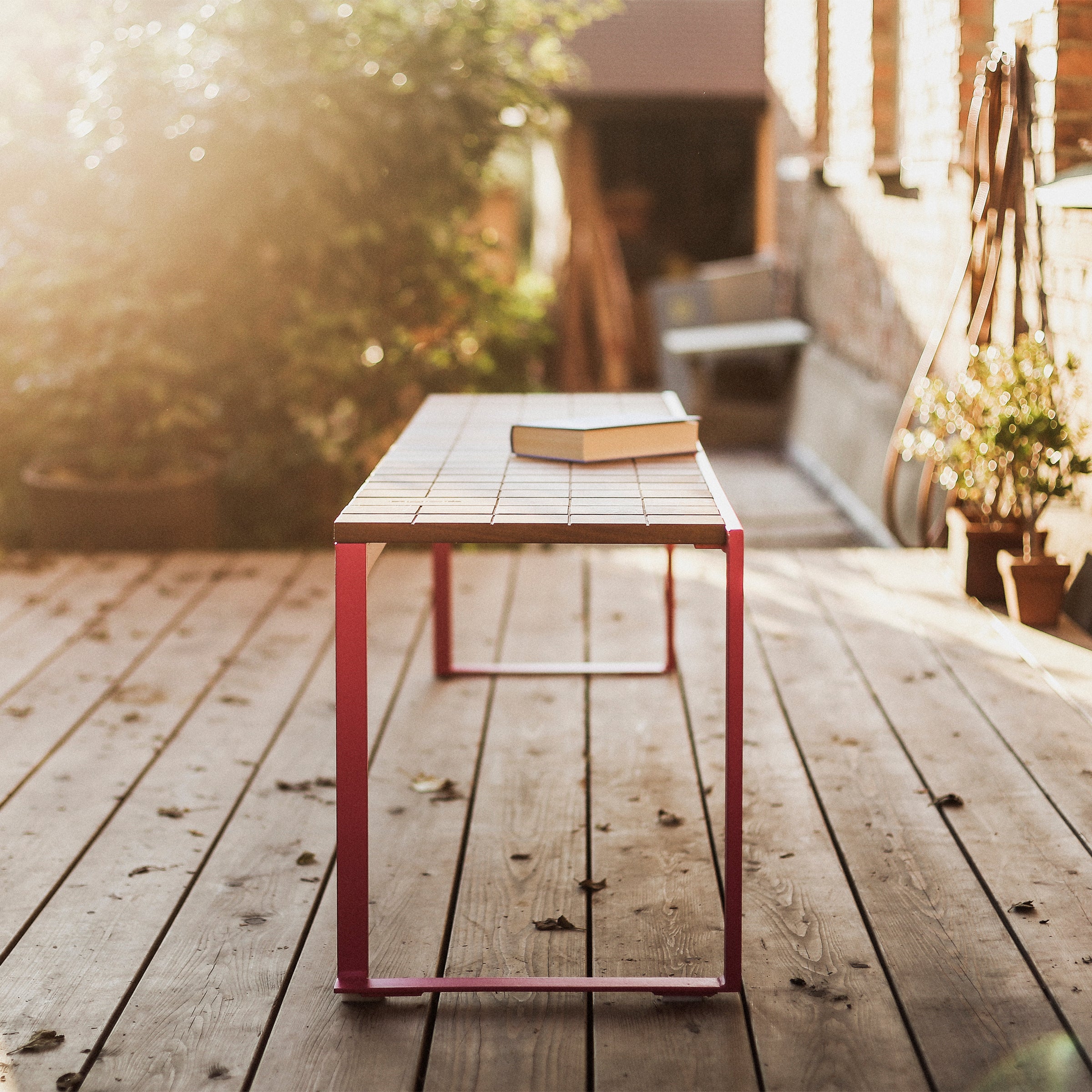 A Bistrot Bench by Egoe, featuring a tropical wood surface and pink zinc-plated steel legs, stands on a sunlit wooden deck with an open book atop. Potted plants and a brick wall form a serene backdrop as sunlight casts warm tones across the scene.