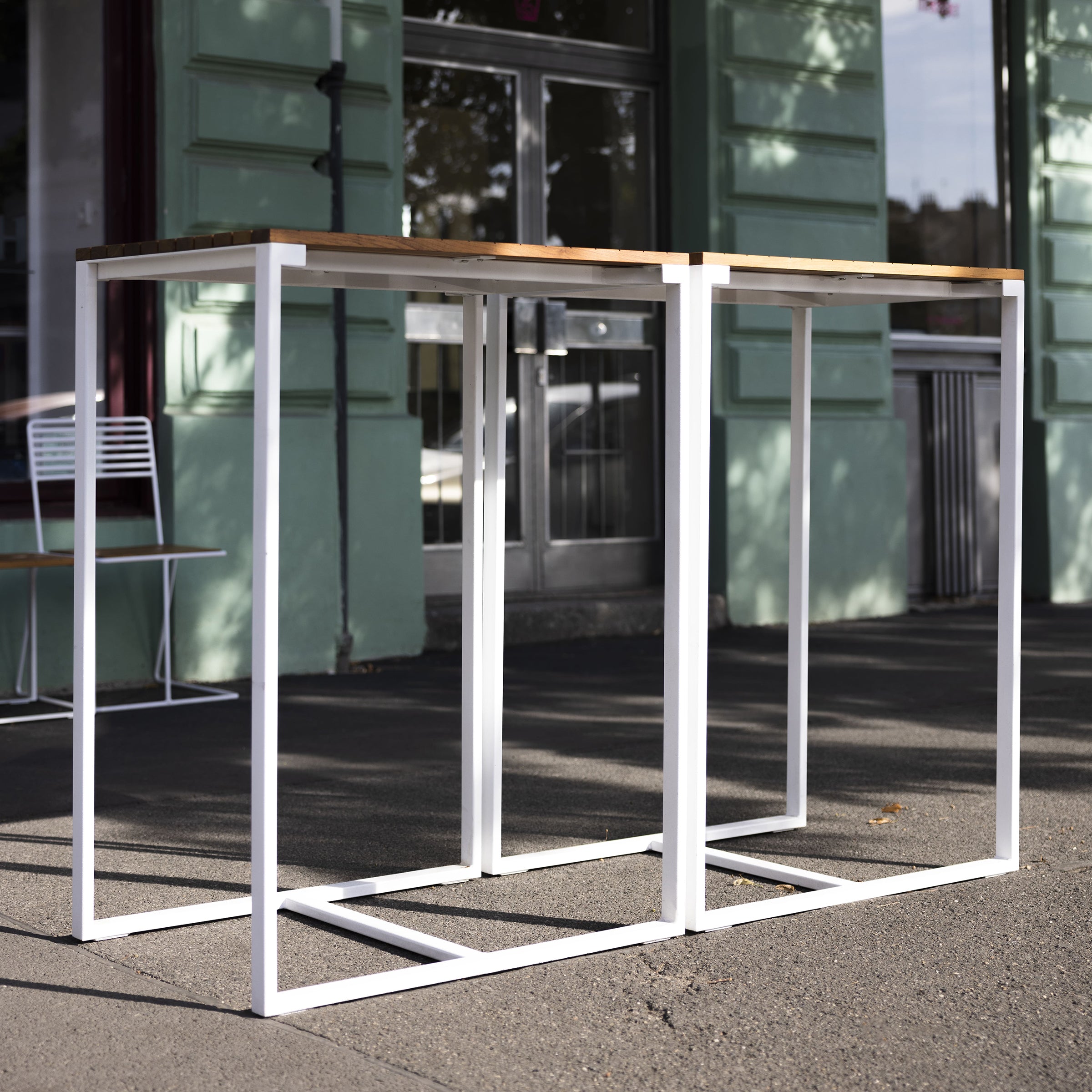 Two Bistrot Tables by Egoe, with white metal frames and tropical wood tops, stand outside a green building beside a large window. A white chair is seen in the background, and shadows of the tables are cast on the sidewalk.