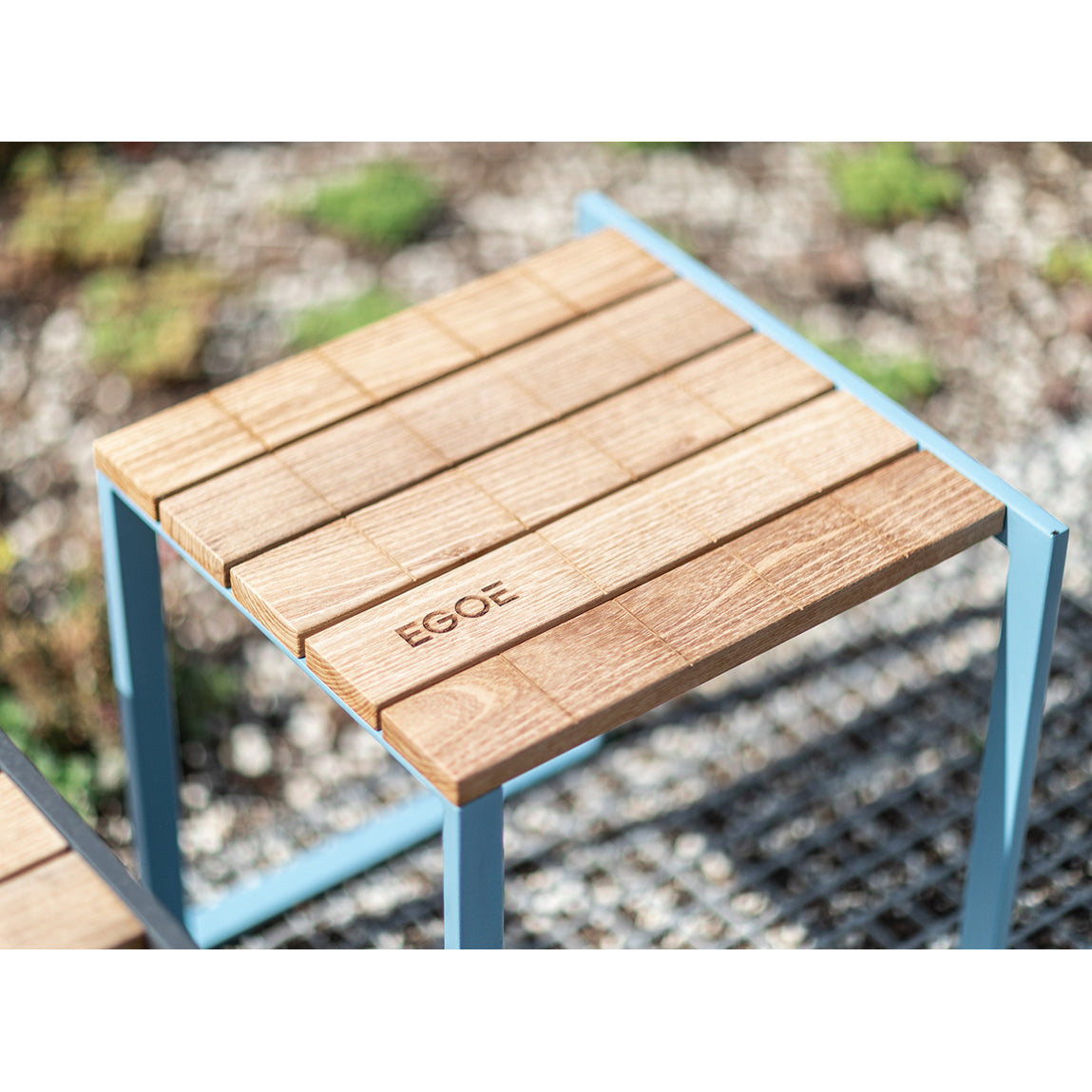Close-up of a wooden outdoor table featuring an Egoe engraving. The tabletop, made with tropical wood, rests on a blue zinc-plated steel frame. A blurred background of gravel and grass enhances the depth of this stylish Bistrot series setup.