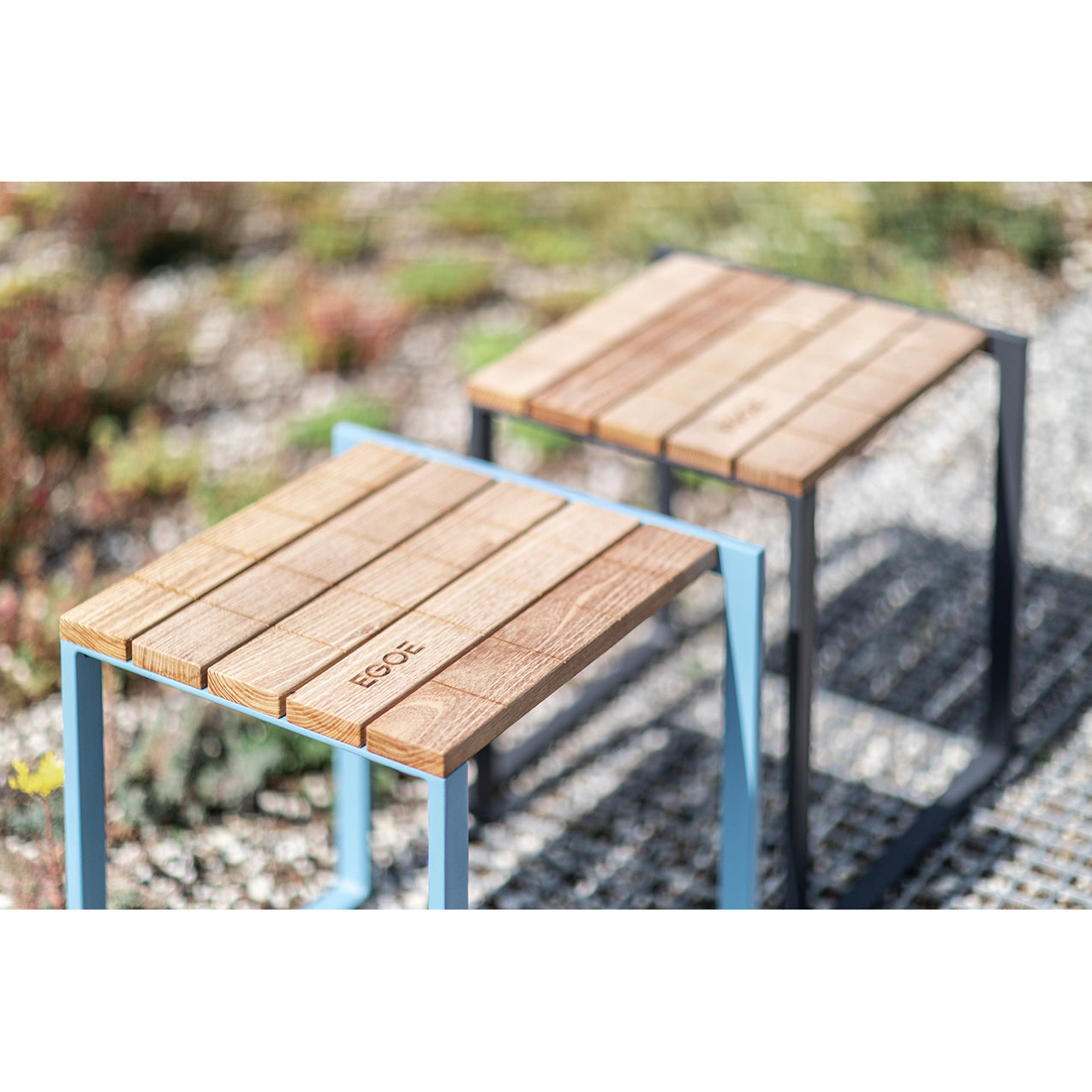 The Egoe Bistrot series features two modern square stools with tropical wood slats and sleek metal frames, ideal for outdoor use on gravel. One stool has engraved text on its top, and both have durable zinc-plated steel foundations, all set against a backdrop of lush plants.