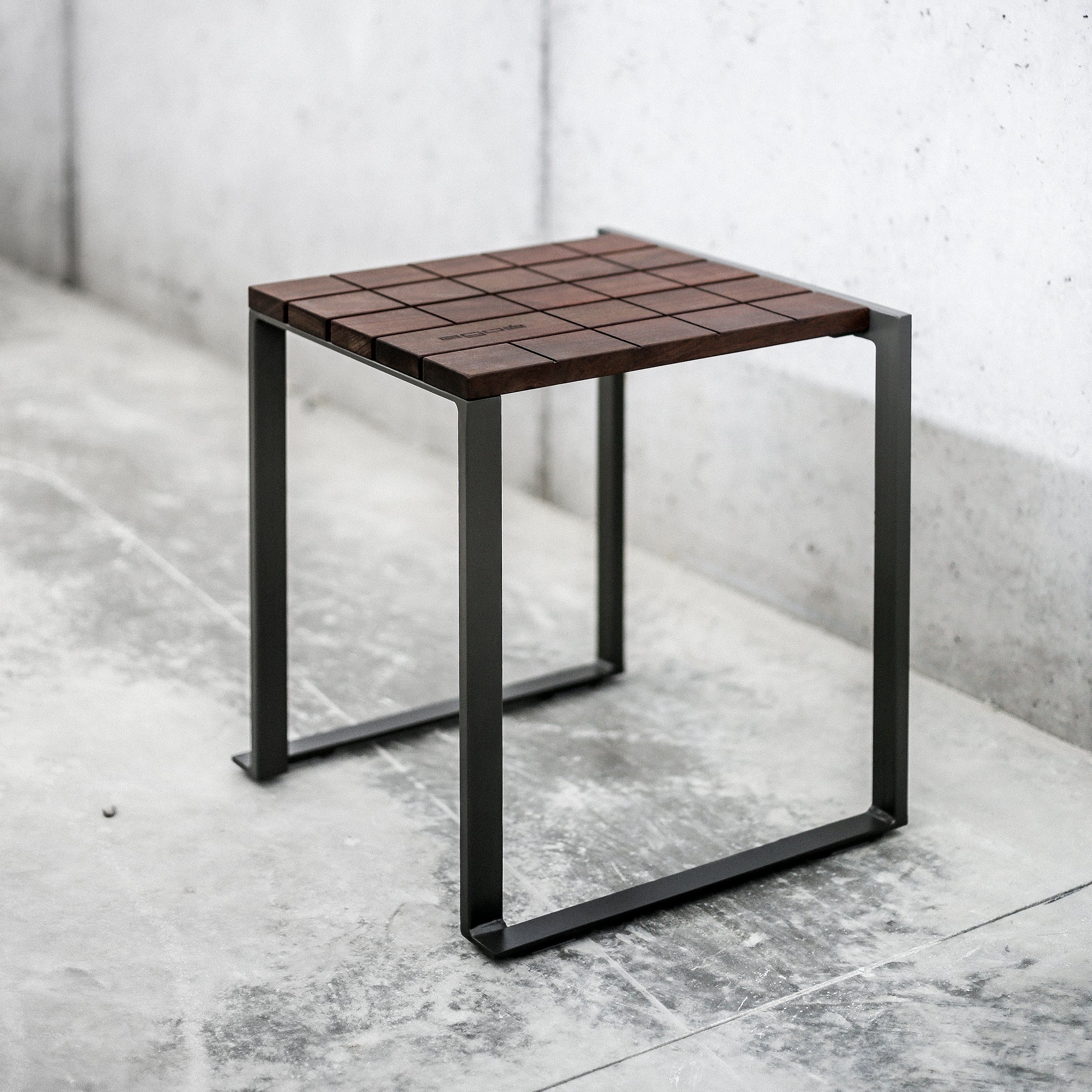 The Bistrot Stool by Egoe features a minimalist design with a square tropical wood seat and powder-coated metal frame, resting on a concrete floor beside a plain light wall. A grid pattern enhances its modern appeal.