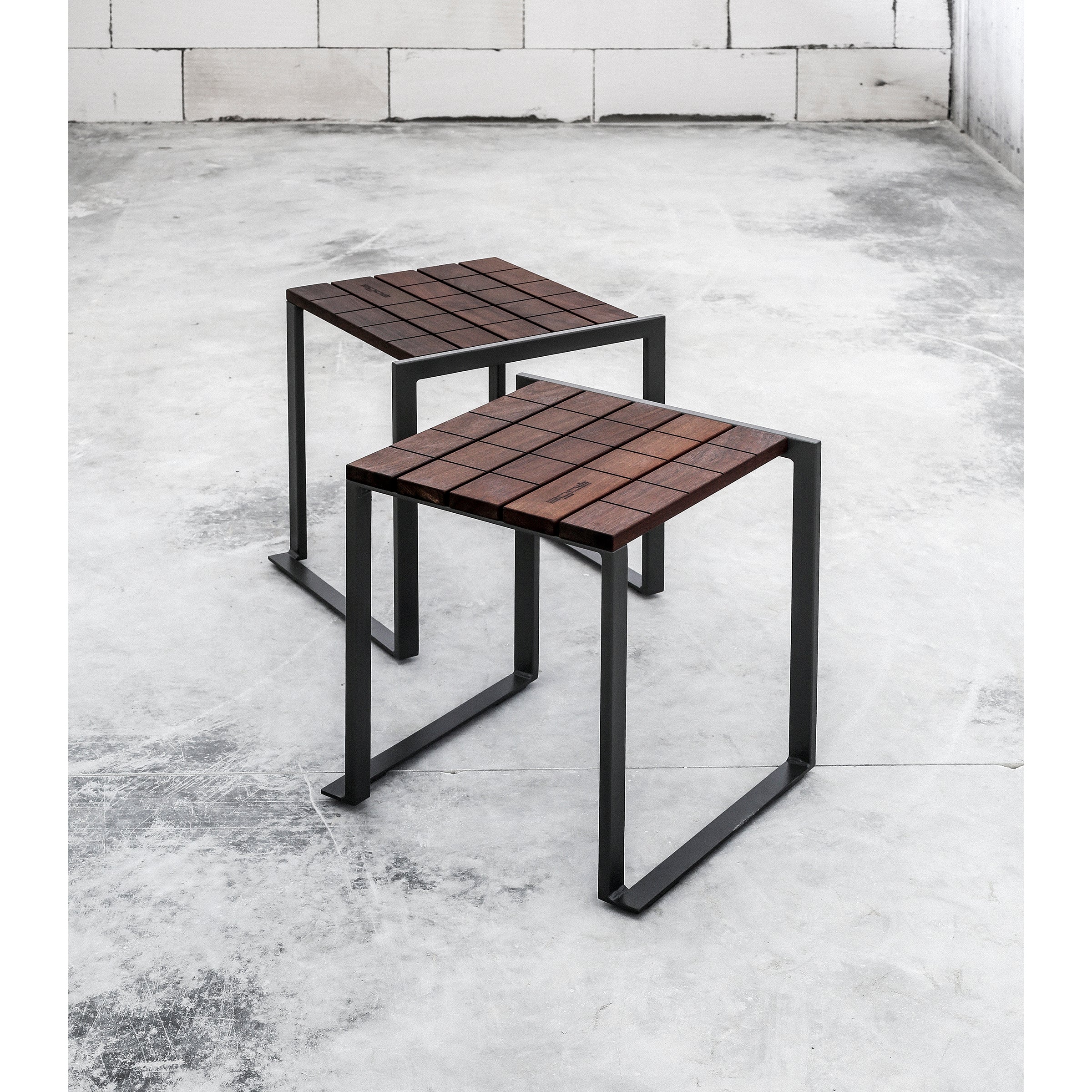 In a stark room with white brick walls, two Egoe Bistrot stools stand on a concrete floor. Their square tropical wood seats contrast elegantly with the black zinc-plated steel frames, angled slightly to add depth to the scene.