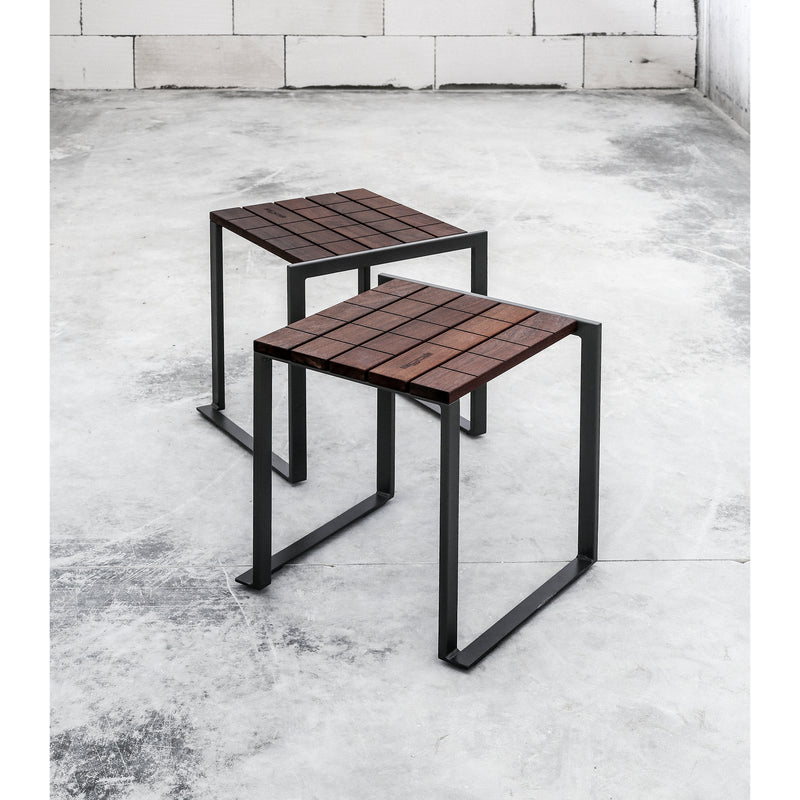 In a stark room with white brick walls, two Egoe Bistrot stools stand on a concrete floor. Their square tropical wood seats contrast elegantly with the black zinc-plated steel frames, angled slightly to add depth to the scene.