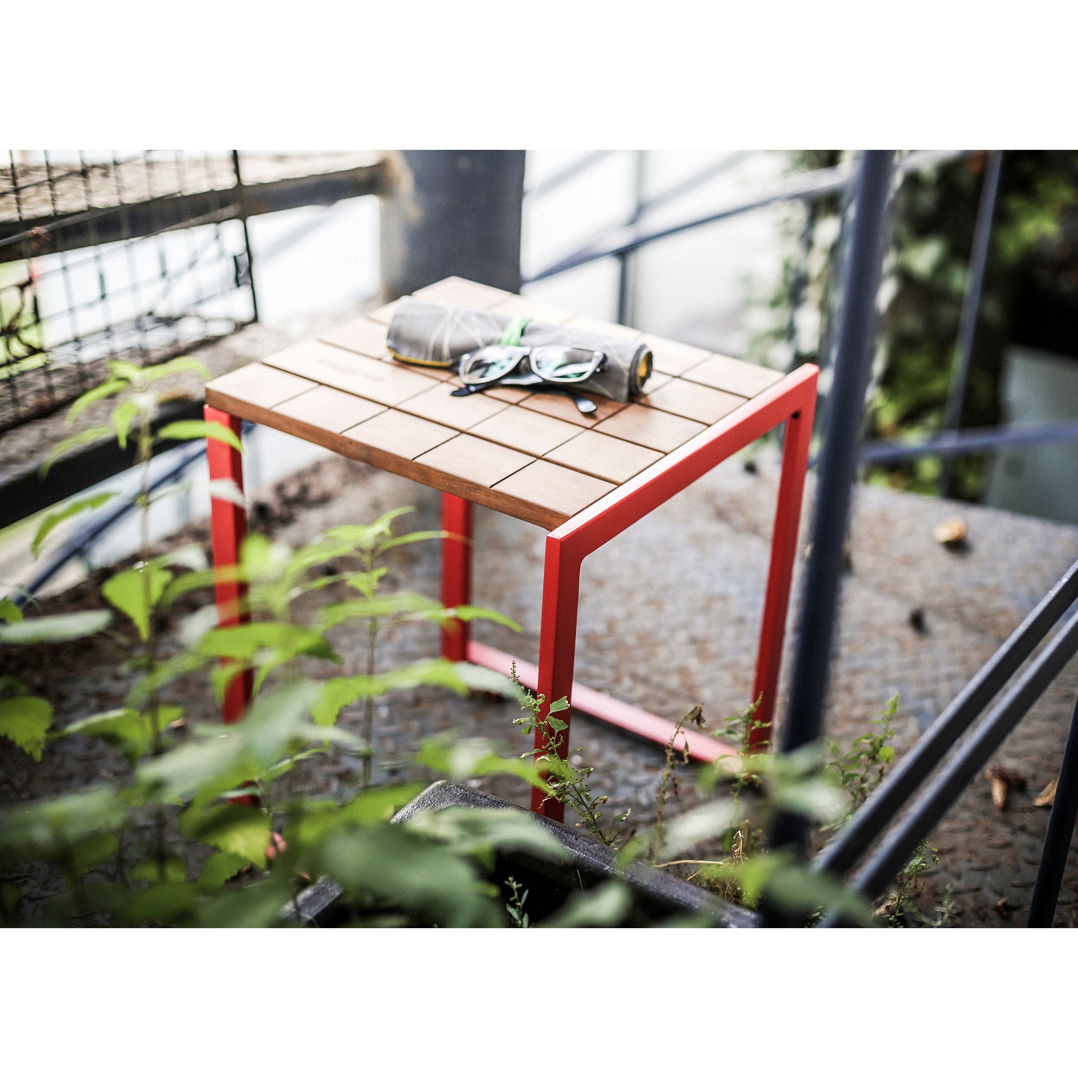 A Bistrot Stool from Egoe, with a red frame and a tropical wood grid-patterned top, sits on a metal stairway. Amid greenery and an industrial backdrop, a pair of sunglasses and a rolled-up magazine rest on it.