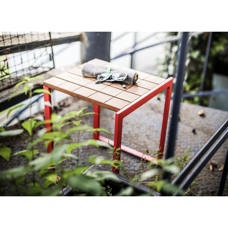A Bistrot Stool from Egoe, with a red frame and a tropical wood grid-patterned top, sits on a metal stairway. Amid greenery and an industrial backdrop, a pair of sunglasses and a rolled-up magazine rest on it.