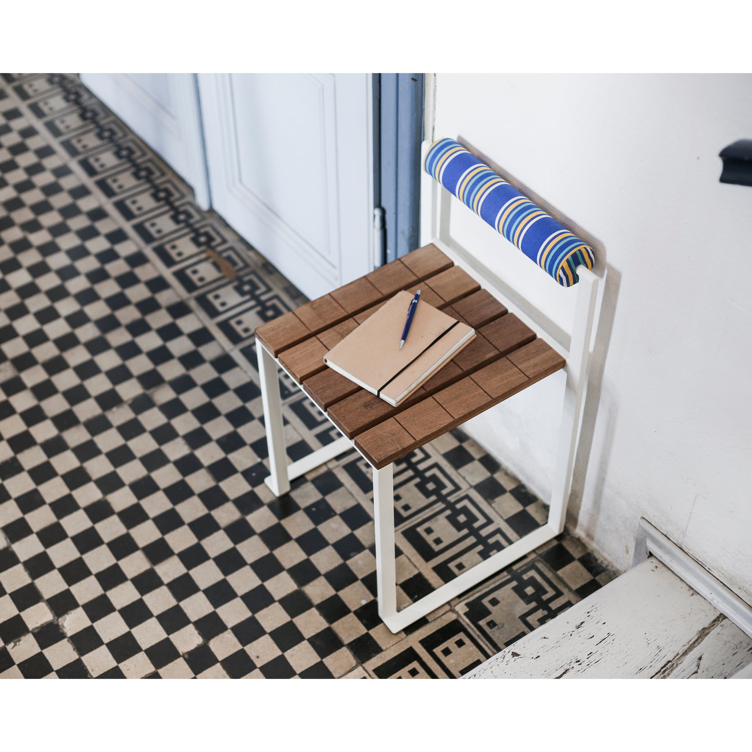 An Egoe Bistrot Stool with Backrest, featuring a zinc-plated steel frame and blue-striped cushion, is placed on black-and-white tile flooring. A notebook and pen lie on the seat against a light-colored wall and closed door in the background.