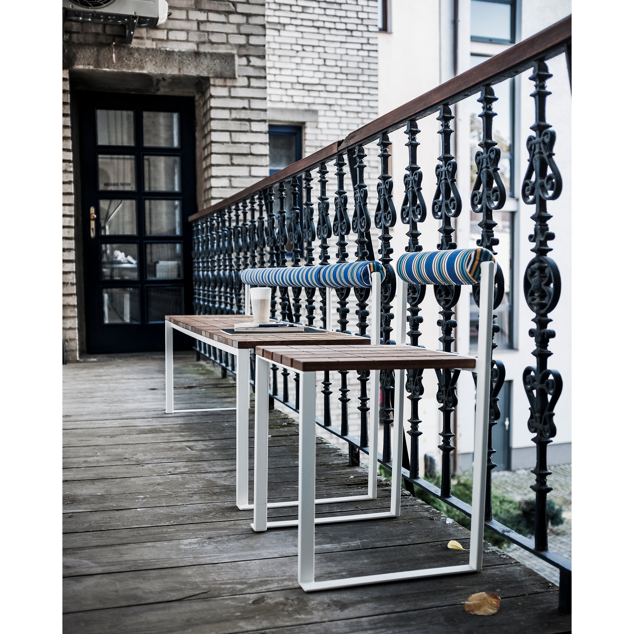 Egoes Bistrot Benches with Backrests, featuring striped cushions, are arranged on a wooden balcony with ornate black railings. A brick building with a black door stands behind them, and a small table holding a cup is placed between the benches.