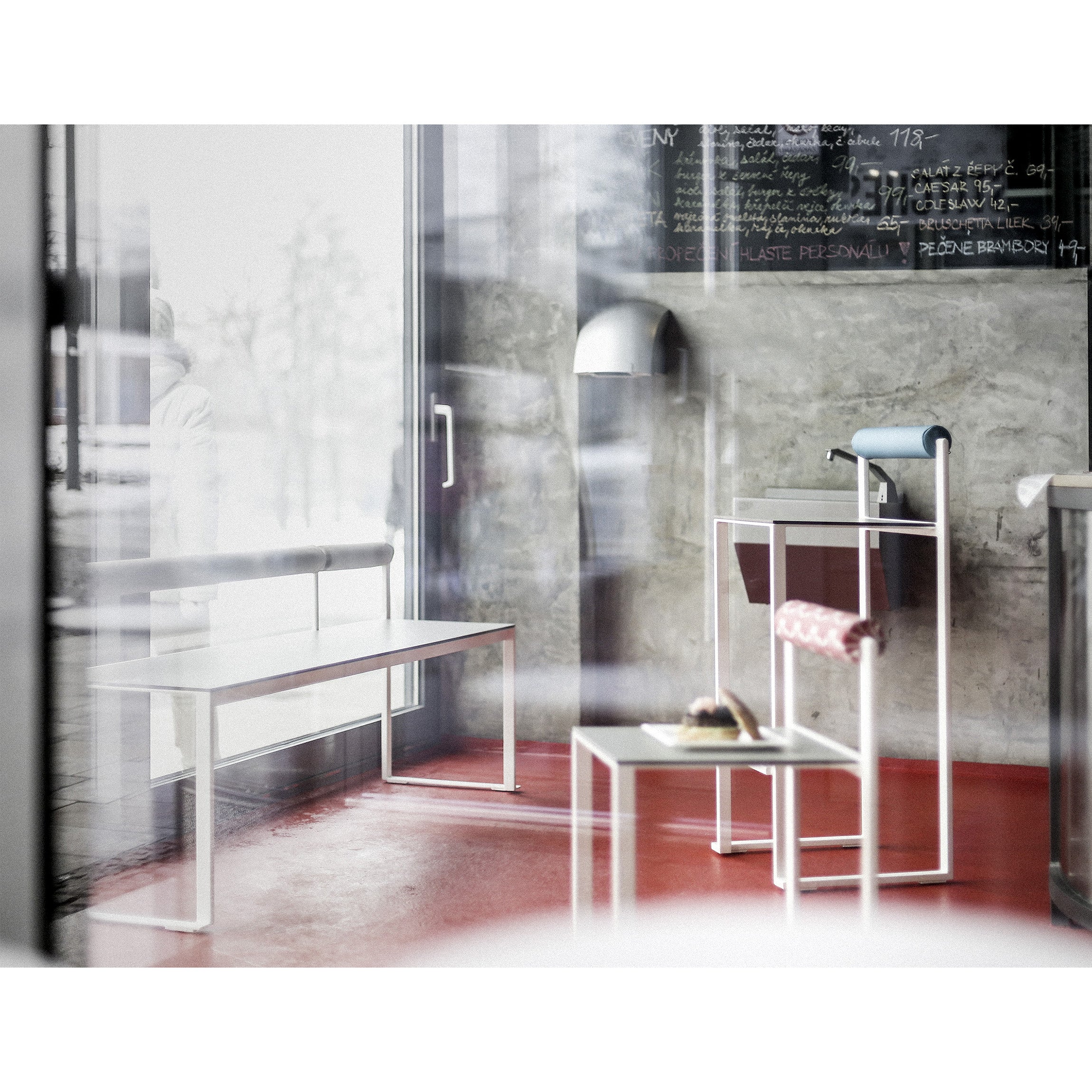 A modern minimalist cafe interior seen through a glass window showcases white Egoe Bistrot benches and a small white table with a pastry on it, red flooring, and a concrete wall partially displaying a chalkboard menu adorned with stackable zinc-plated steel Egoe Bistrot Barstools with Backrest.