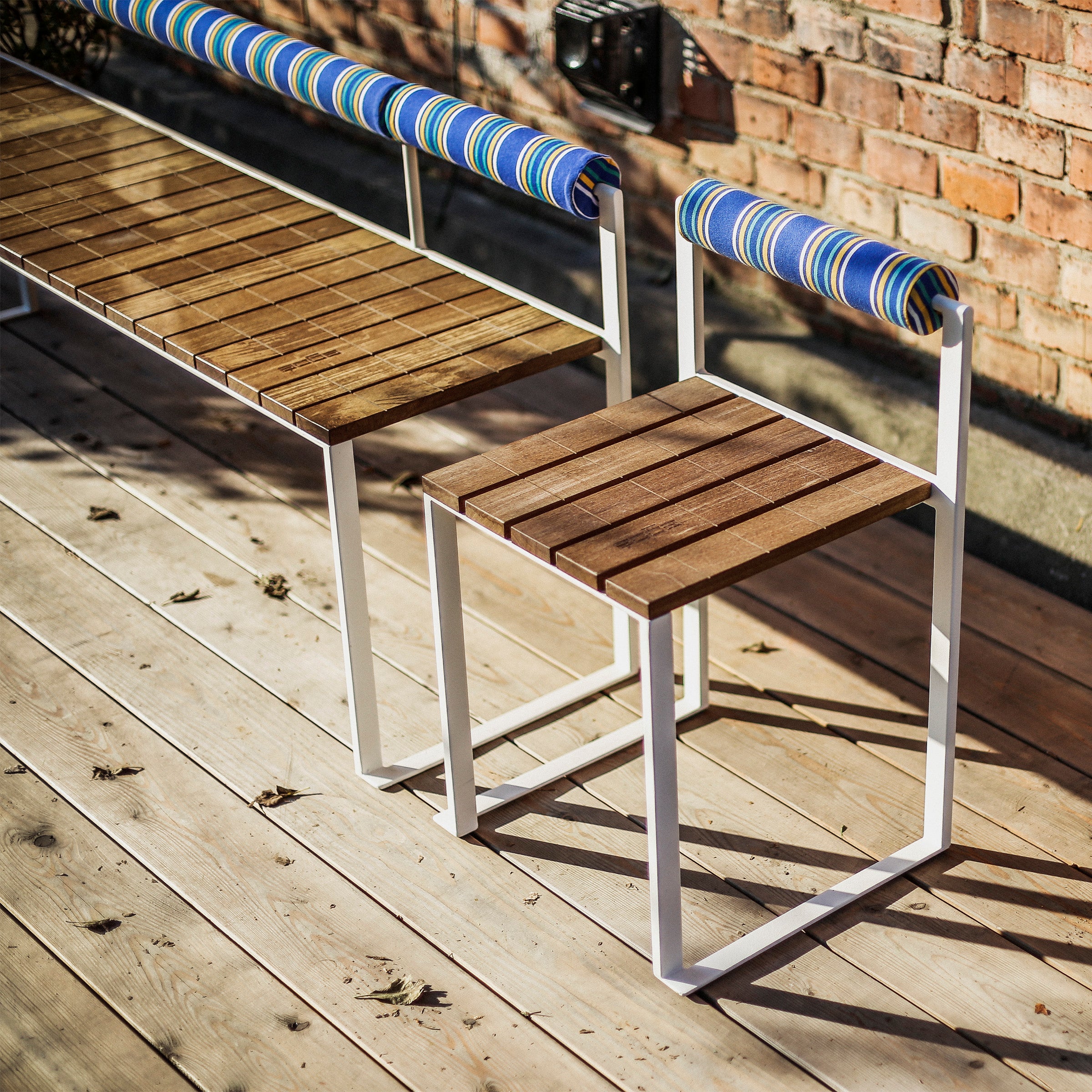The Egoe Bistrot Bench with Backrest features white zinc-plated steel frames and blue striped cushions, beautifully showcased on a sunlit wooden deck against a brick wall with scattered leaves.