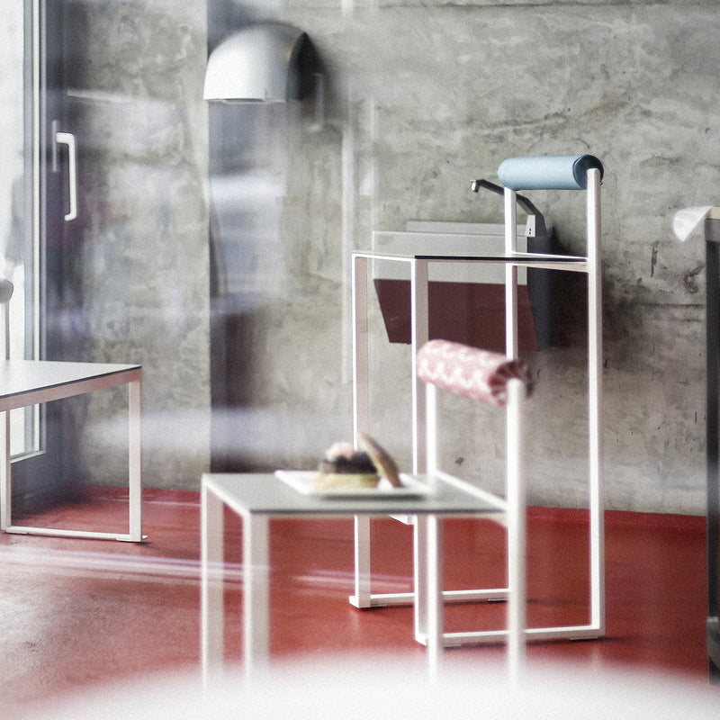 A modern café interior features a concrete wall, red floor, and glass entrance. A small table with a pastry, minimalist chairs from Egoes Bistrot series, an industrial sink with a blue towel, and bright lighting highlighting Bistrot Barstools with Backrest create reflections on the glass surface.