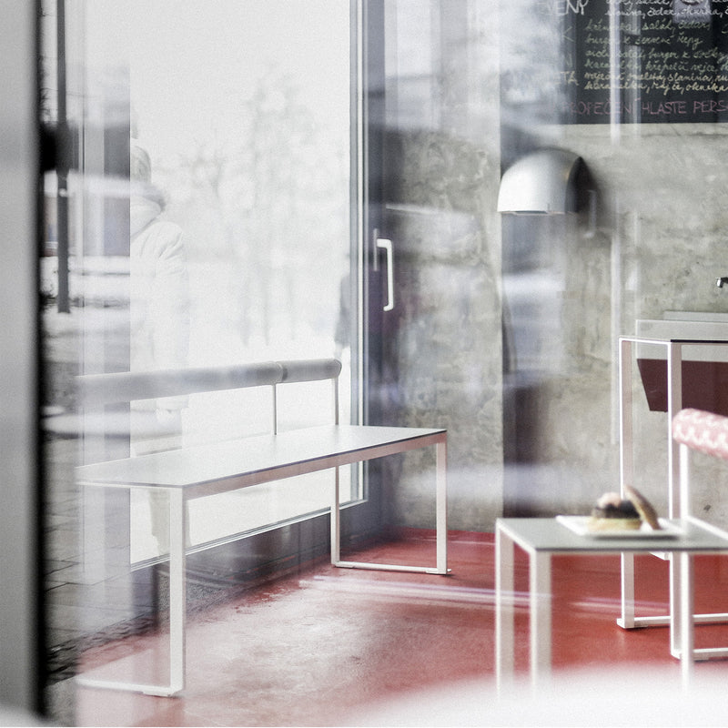 The modern café features a spacious, cozy interior with a red floor and white furniture from Egoes Bistrot series, including the Bistrot Bench with Backrest placed against a glass wall. Large windows flood the space with natural light, highlighting minimalistic design enhanced by tropical wood accents.