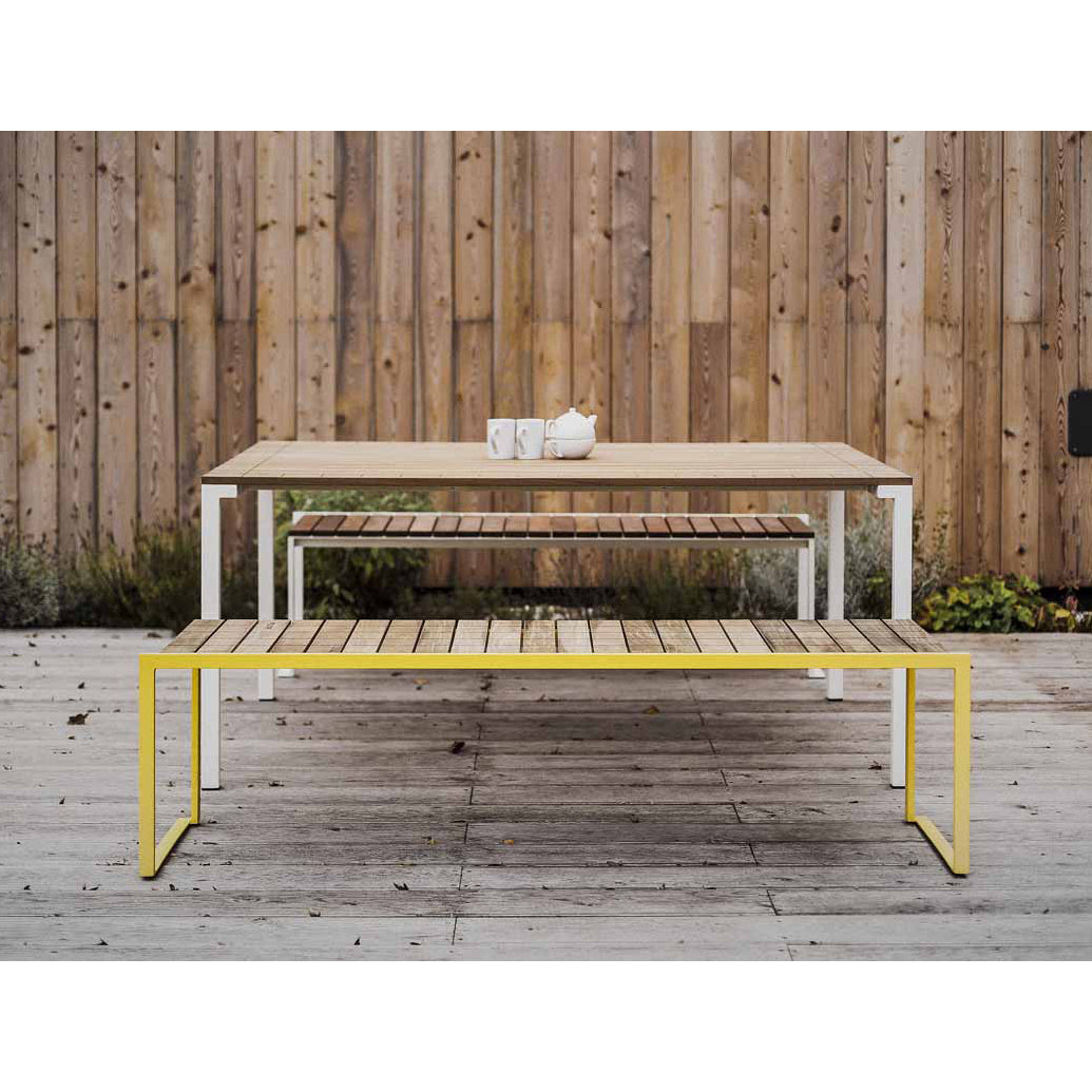 The Bistrot Table by Egoe is an outdoor wooden dining set with three tables of varying sizes, featuring minimalist zinc-plated steel frames. The largest table has a white frame, the smallest a yellow frame, and together they provide tropical wood elegance on a paved ground against a wooden fence.