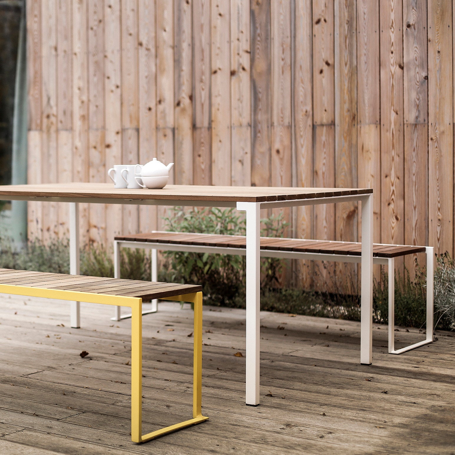 The Bistrot Table by Egoe features a wooden table with white zinc-plated steel legs, complemented by matching benches (one in vibrant yellow). The set is adorned with a white teapot and cups on top, all set against a wooden-paneled wall.
