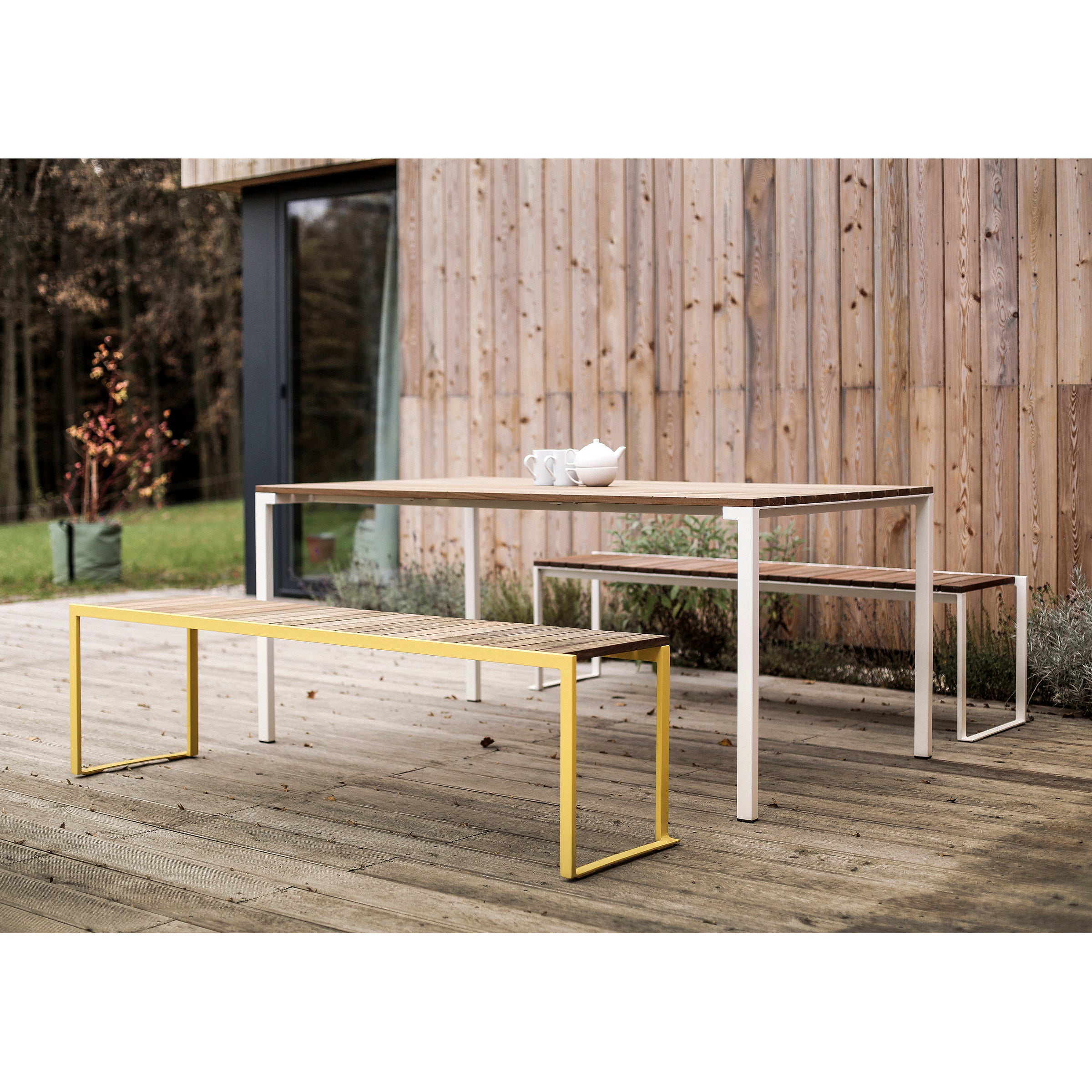 The outdoor patio features an Egoe Bistrot Table set with two benches, one white and the other yellow. In the background are a wooden building and door, with trees and a potted plant. Zinc-plated steel accents enhance its modern charm.