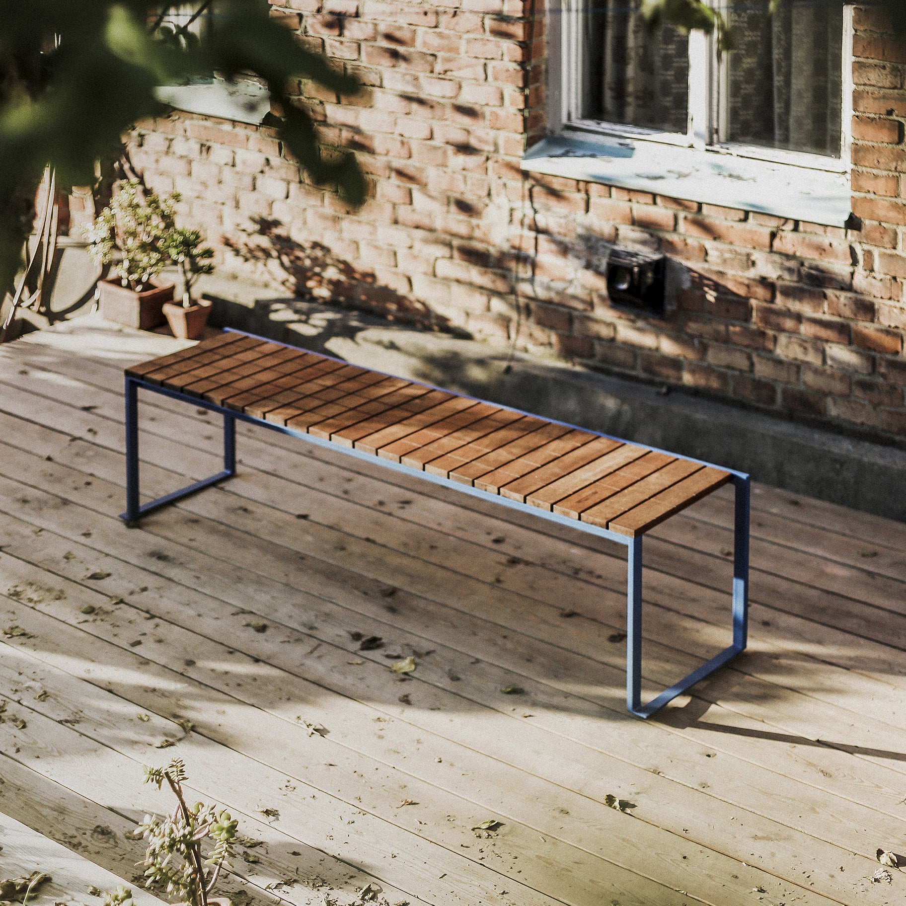A sleek Egoe Bistrot Bench, crafted from tropical wood with a zinc-plated steel frame, sits on a sunlit deck. A brick wall with a window and potted plants is in the background, creating a bright and tranquil scene with soft shadows from surrounding foliage.