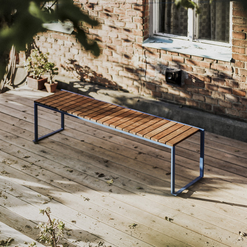 A sleek Egoe Bistrot Bench, crafted from tropical wood with a zinc-plated steel frame, sits on a sunlit deck. A brick wall with a window and potted plants is in the background, creating a bright and tranquil scene with soft shadows from surrounding foliage.