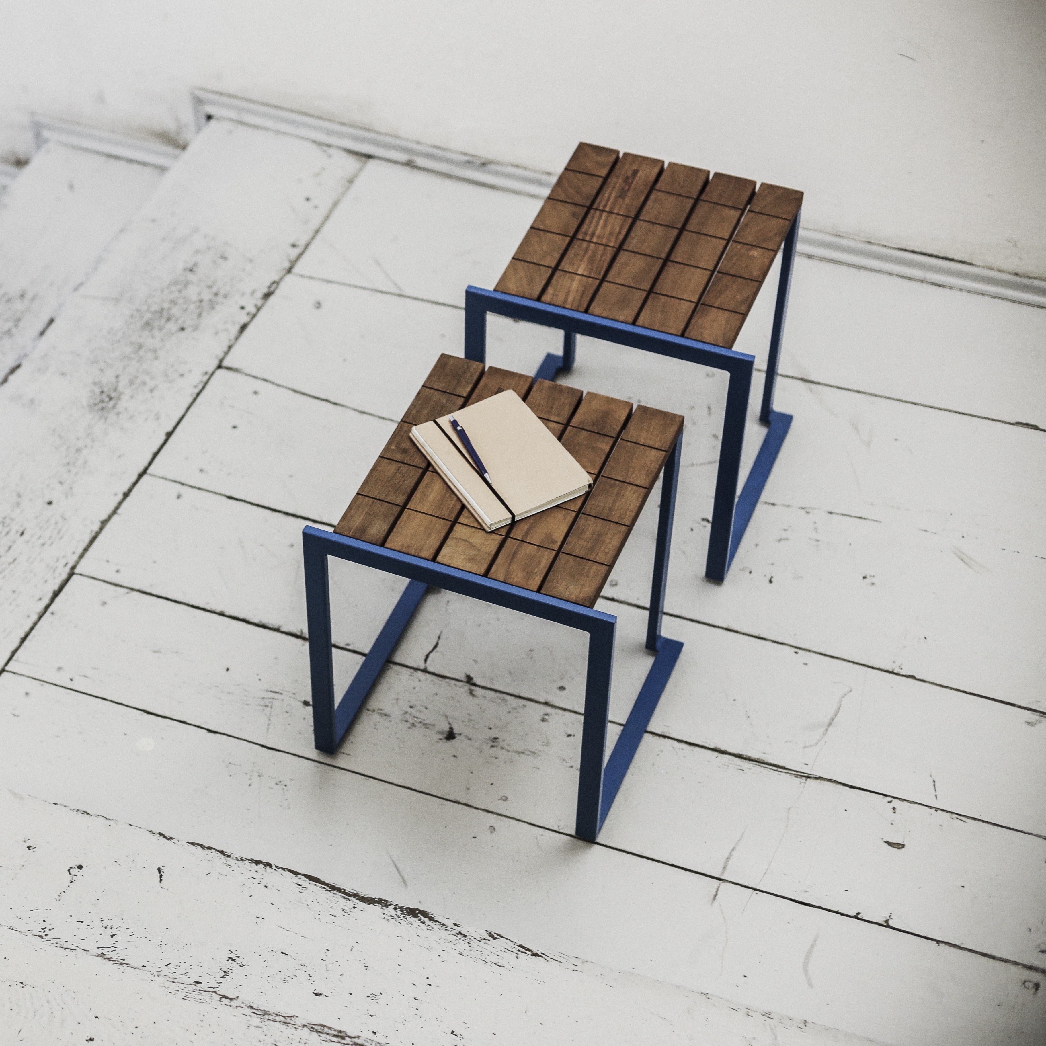 Two Bistrot stools by Egoe, with blue metal frames and slatted tropical wood tops, stand on a weathered white floor. One holds a closed notebook and pen.