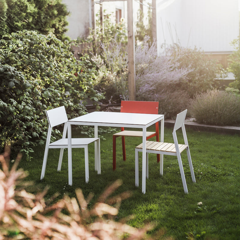 The Cora Table by Egoe, part of a chic outdoor dining set, includes a white table and three modern chairs—two white and one red—all made of powder-coated aluminum. This contemporary ensemble looks stunning on a green lawn amidst lush greenery in gentle sunlight.