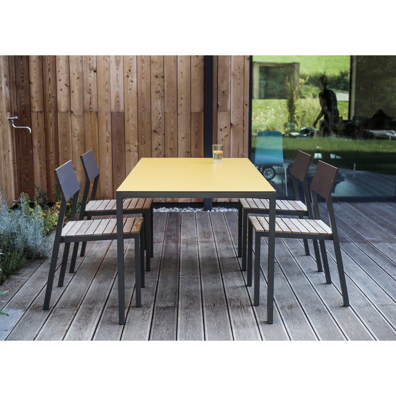 An outdoor dining space includes an Egoe Cora Table, in yellow and rectangular, with five metal-framed chairs featuring wooden seats on a wooden deck. Powder-coated aluminum accents enhance the elegance, while a glass of water rests on the table, reflecting garden views.