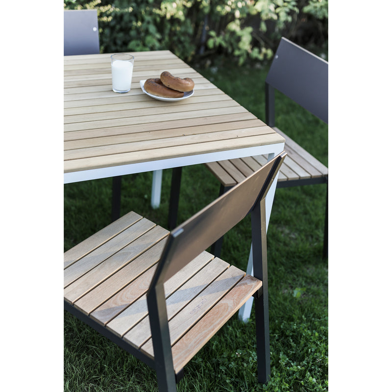 The Egoe Cora Table, paired with four powder-coated aluminum chairs, complements a grassy lawn. A glass of milk and two hot dogs on a plate add to this contemporary setup, nestled in a serene garden with trees or bushes in the background.