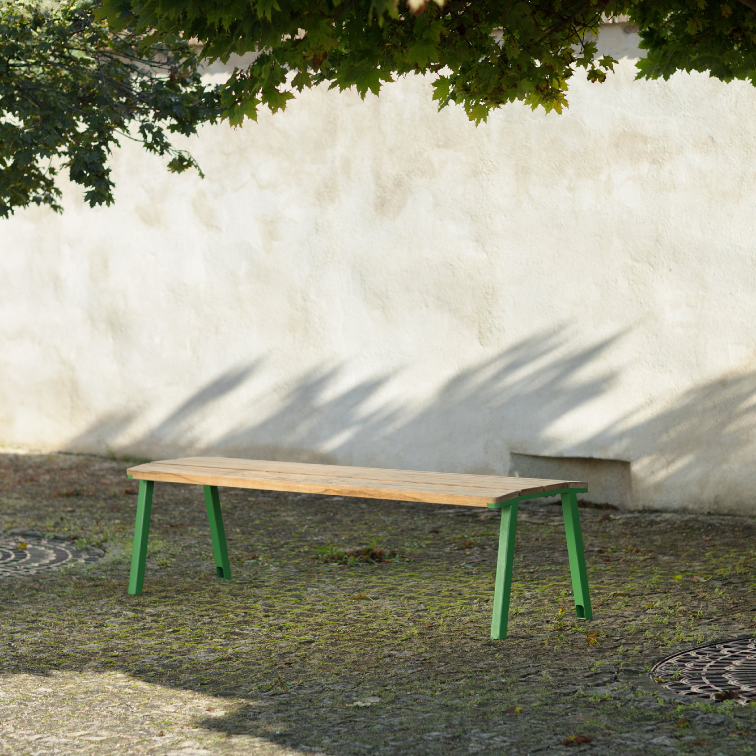 The Cora Public Bench by Egoé, crafted from tropical Jatoba wood with green legs, sits on a cobblestone path against a white wall. Overhead tree branches cast leafy shadows, enhancing the calm outdoor setting with Egoé’s street furniture elegance.