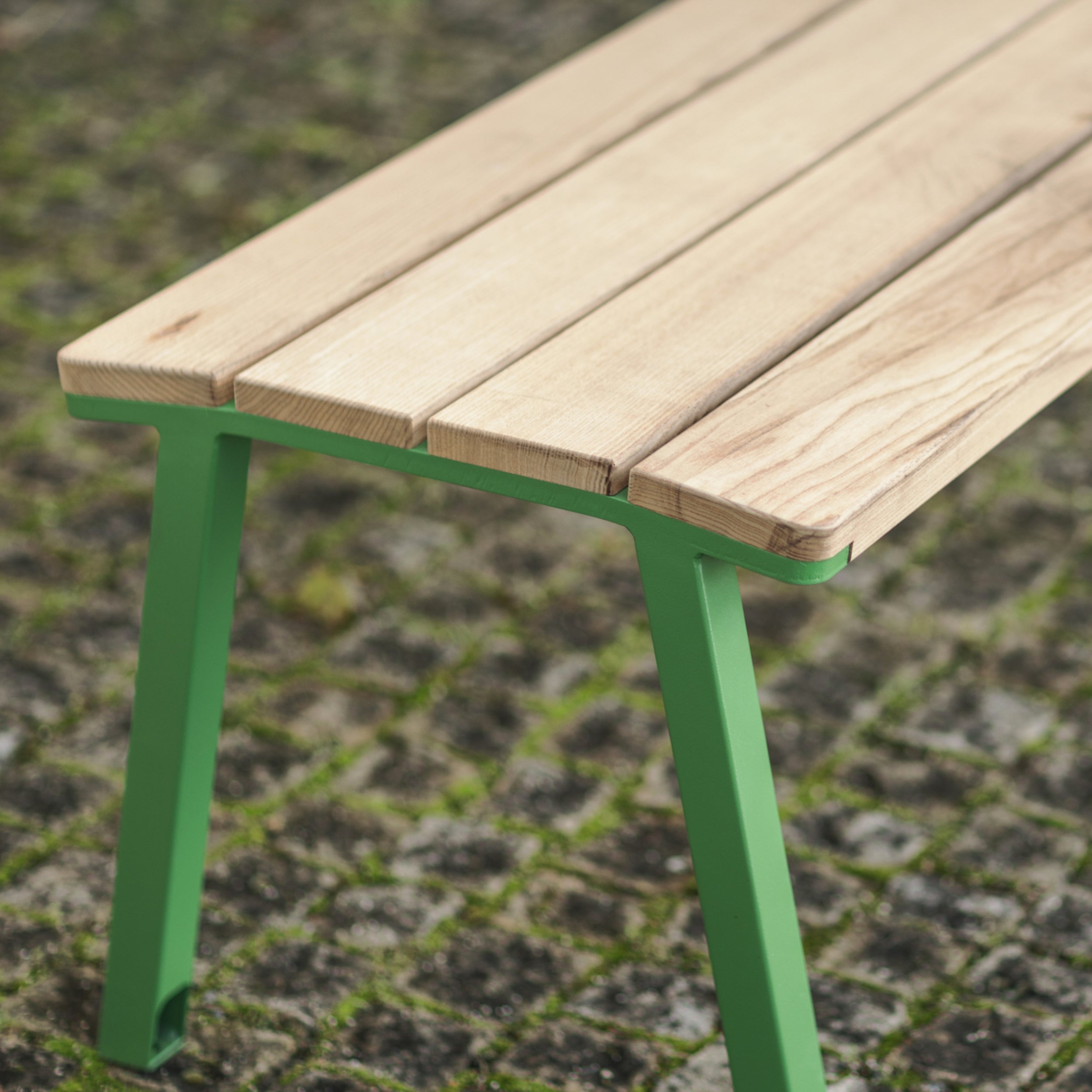 The Cora Public Bench by Egoe, featuring a wooden seat made from tropical Jatoba wood and green metal legs of zinc and powder-coated steel, sits on a mossy cobblestone ground.