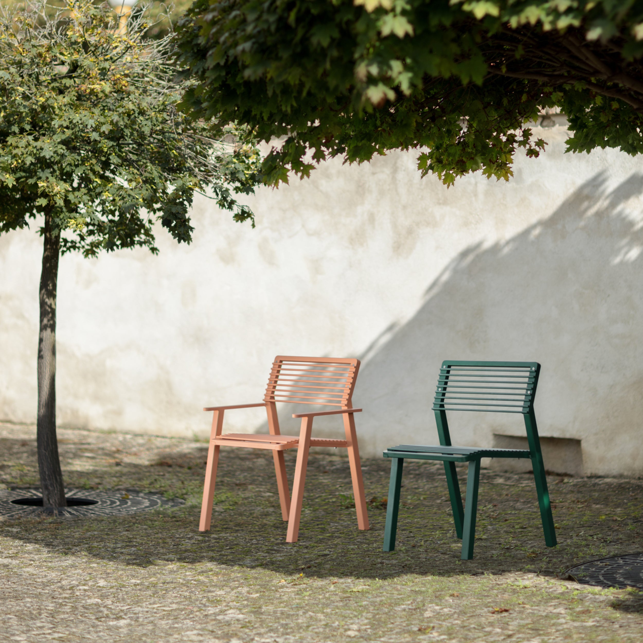 Two empty chairs, one orange and one green, from the Egoé Cora Public Chair collection, are placed under a leafy tree against a light-colored wall. Crafted from tropical Jatoba wood, these pieces rest on cobblestones, casting long shadows.