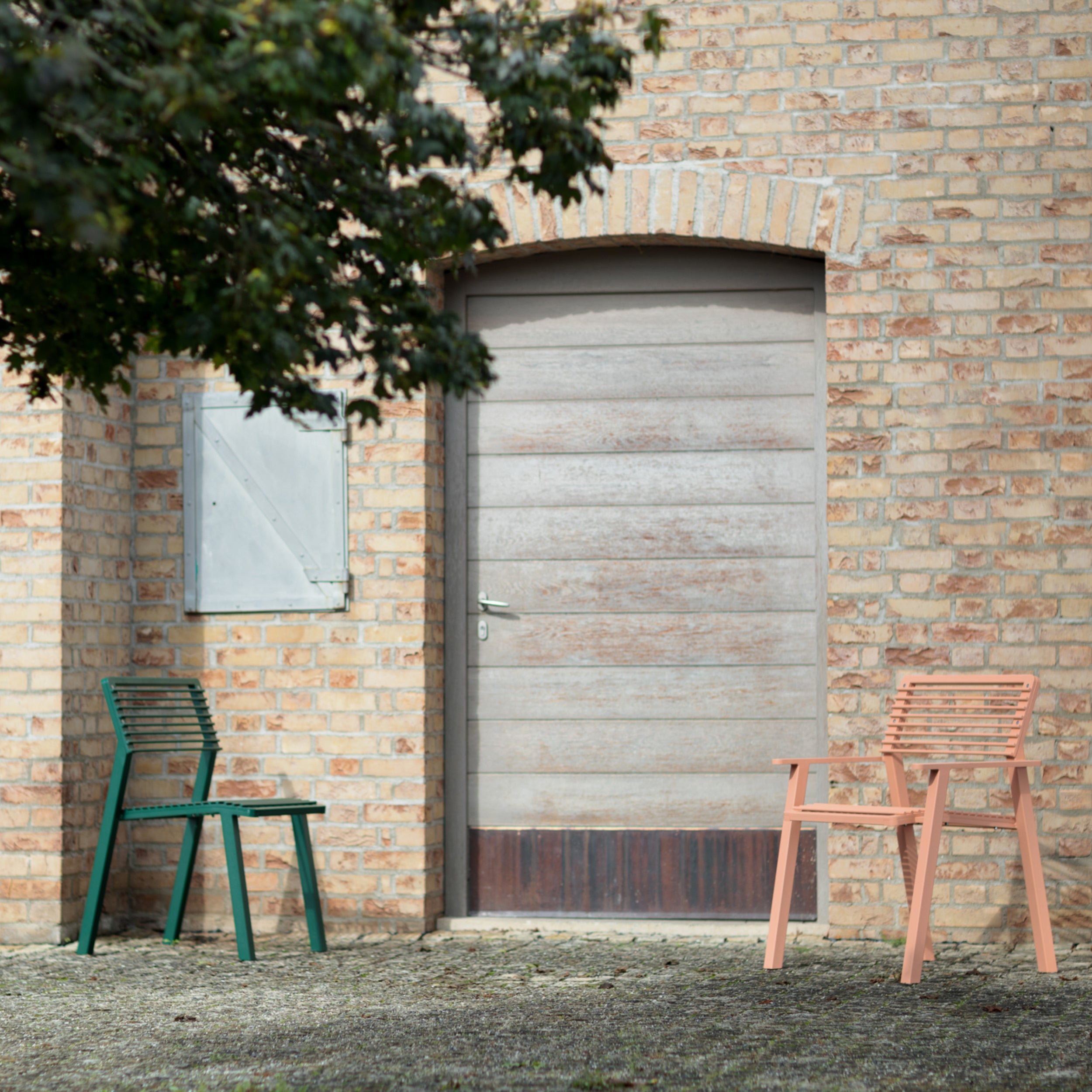 Two Cora Public Chairs by Egoe, crafted from tropical Jatoba wood, one green and one peach, are positioned near a rustic brick wall with a closed wooden door. A leafy tree partially shades the scene, casting soft shadows on the ground.
