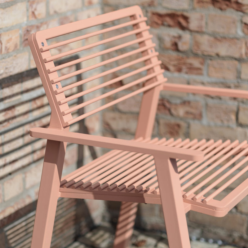 Close-up of a peach-colored Cora Public Chair by Egoe on a brick patio. This street furniture piece has a minimalist design, with parallel slats casting shadows on the ground. Sunlit bricks provide a textured backdrop.