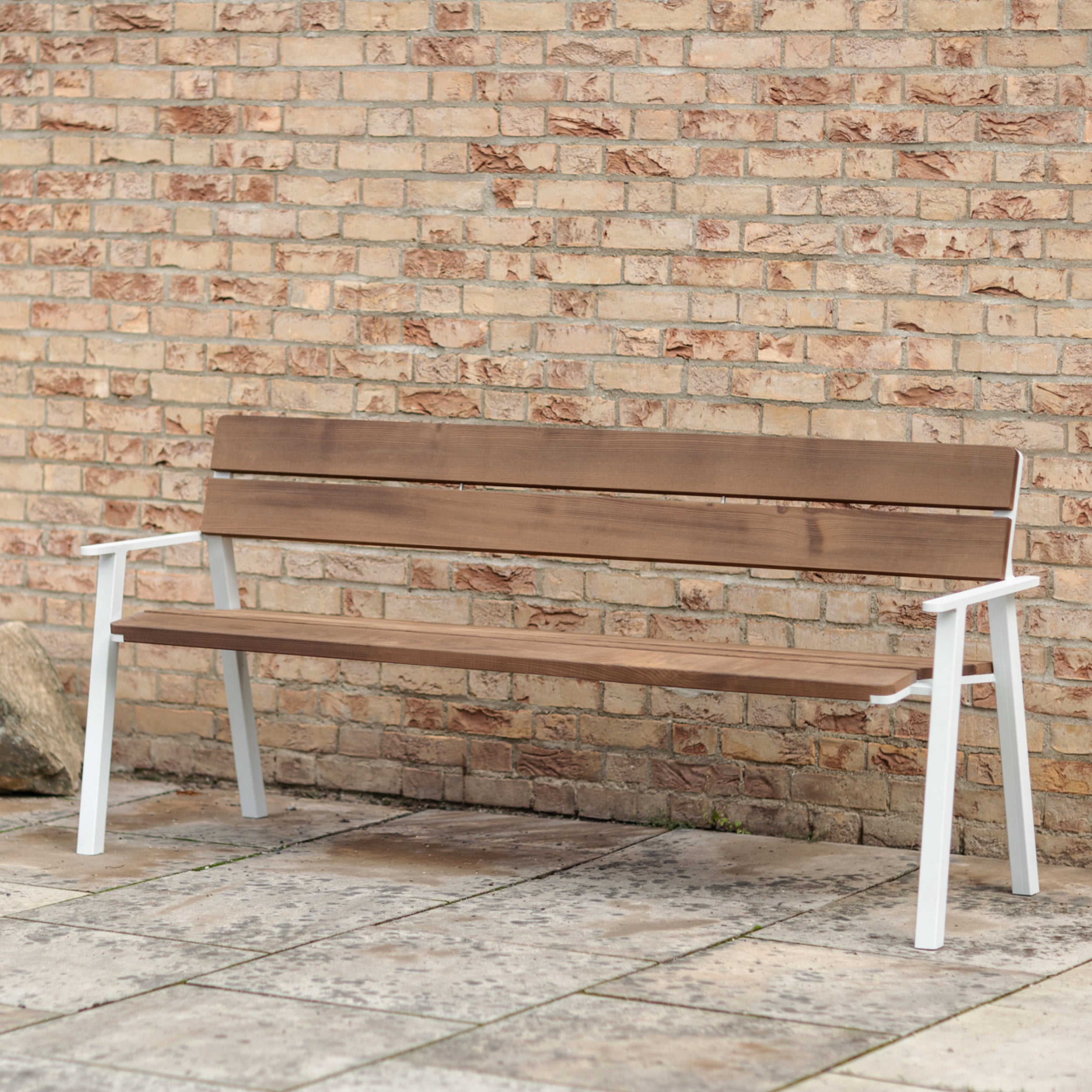The Cora Public Bench by Egoé, featuring white metal legs and armrests combined with tropical Jatoba wood, stands on a stone-paved area against a beige brick wall. Its minimalist design and natural finish offer a simple and serene setting.